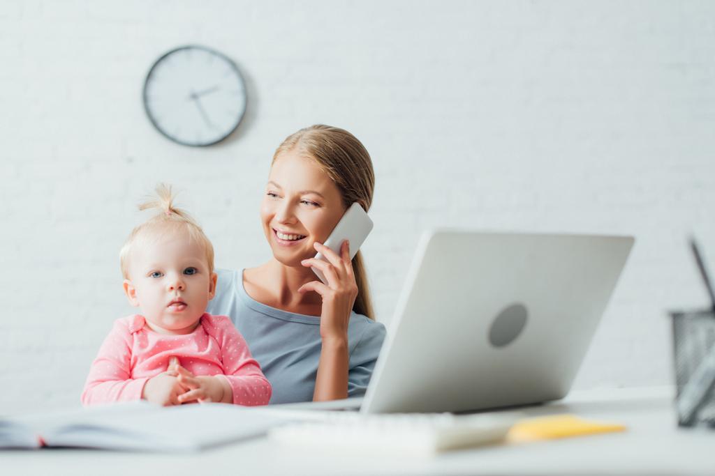 Enfoque selectivo de la mujer hablando en el teléfono inteligente mientras trabaja cerca de la hija bebé en casa  - Foto, Imagen