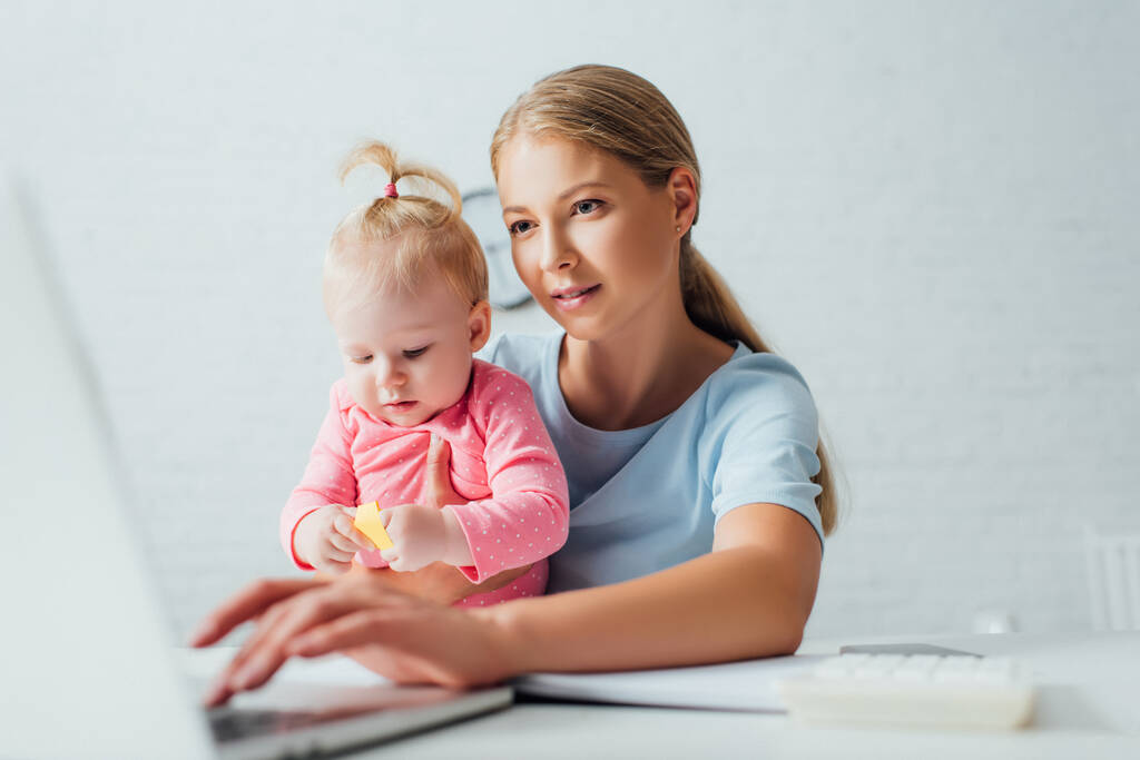 Concentration sélective de pigiste en utilisant un ordinateur portable tout en tenant bébé fille à table  - Photo, image