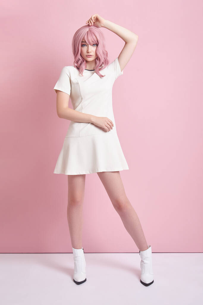 Hermosa chica con el pelo rosa, coloración del pelo. Mujer linda anime se encuentra sobre un fondo rosa en un vestido blanco corto. Cabello teñido, peinado perfecto - Foto, Imagen