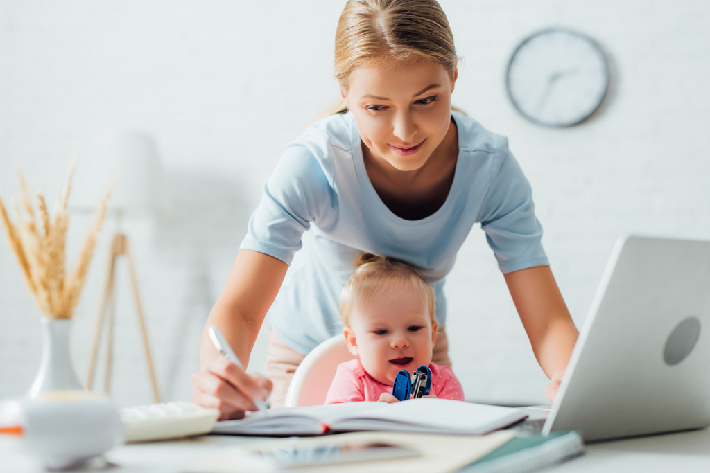 A jegyzetfüzetbe író nő szelektív fókusza, miközben a csecsemő közelében dolgozik tűzőgéppel az asztalnál  - Fotó, kép