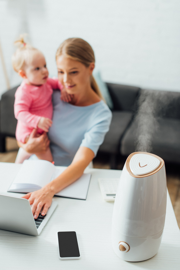 Selectieve focus van luchtbevochtiger op tafel en moeder houden van baby meisje tijdens het gebruik van laptop thuis  - Foto, afbeelding