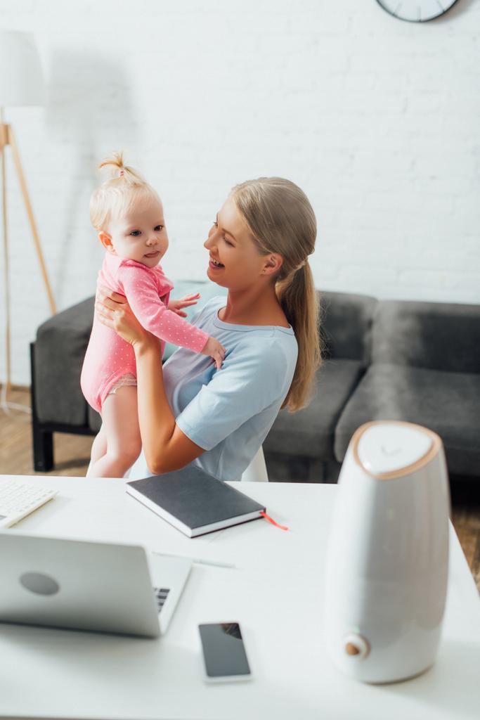 Selective focus of mother holding baby girl near gadgets, notebook and humidifier on table  - Photo, Image