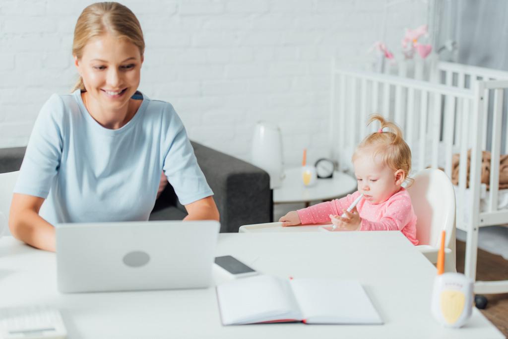 Enfoque selectivo de la niña sosteniendo la pluma mientras la madre usa el ordenador portátil en casa  - Foto, Imagen
