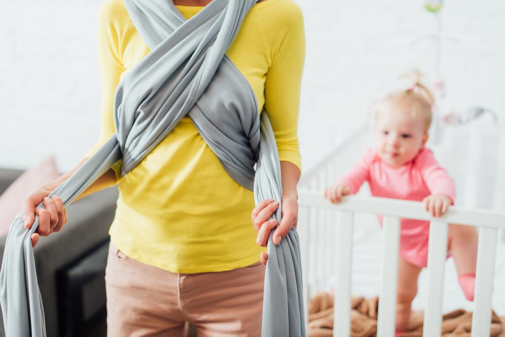 Enfoque selectivo de la mujer que usa cabestrillo de bebé cerca de la hija en cuna  - Foto, Imagen