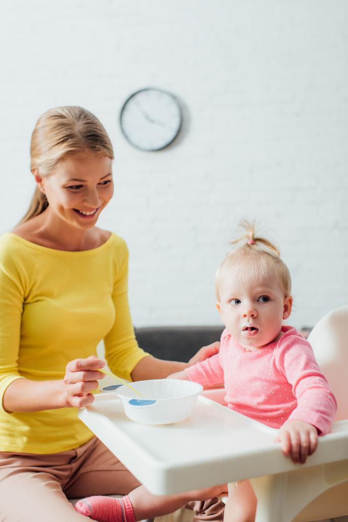 Concentration sélective de la mère regardant bébé fille près du bol et cuillère sur la chaise d'alimentation  - Photo, image