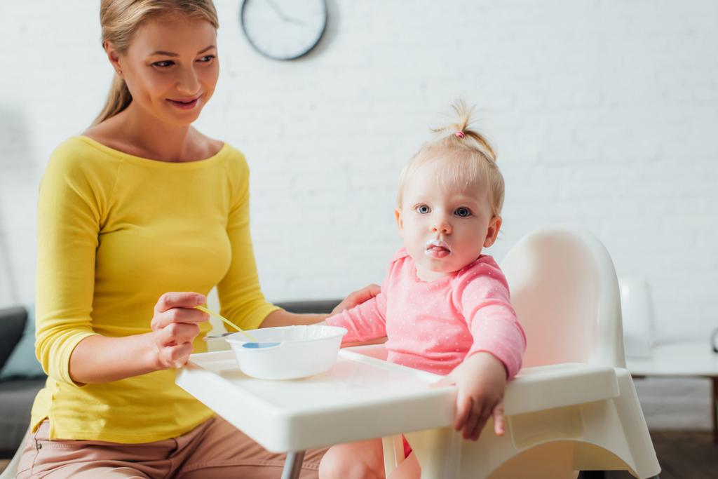 Enfoque selectivo de la niña mirando a la cámara cerca de la madre con cuchara y tazón de comida para bebés en casa  - Foto, Imagen