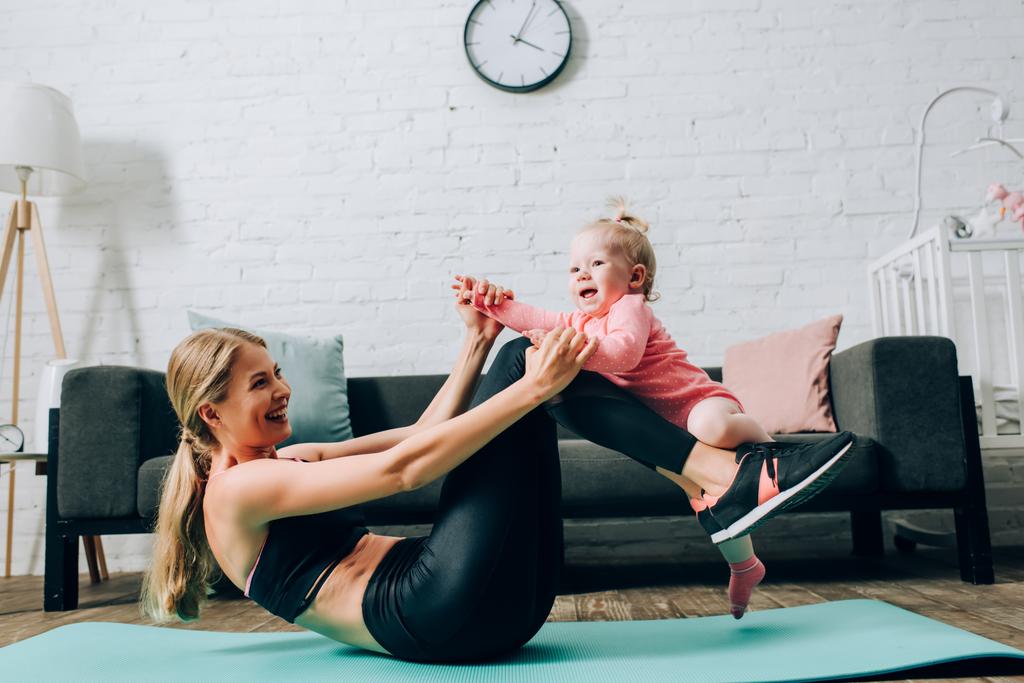Donna in abbigliamento sportivo che tiene in braccio la figlia neonata mentre si esercita sul tappeto fitness in soggiorno  - Foto, immagini
