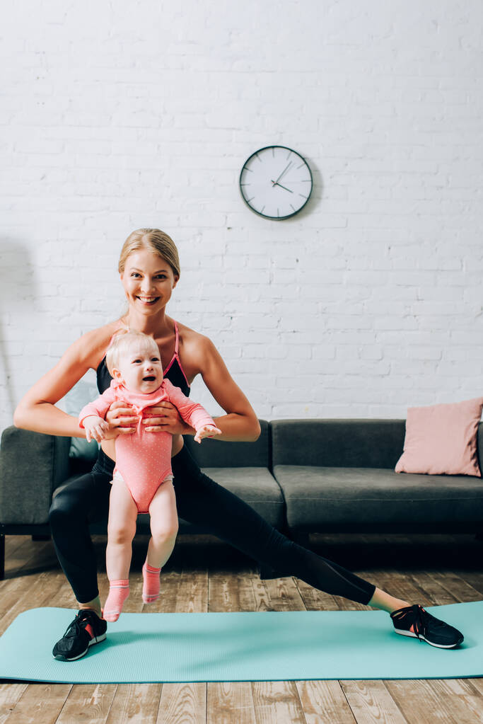 Sportvrouw doet lunges terwijl het houden van baby meisje op fitness mat thuis  - Foto, afbeelding