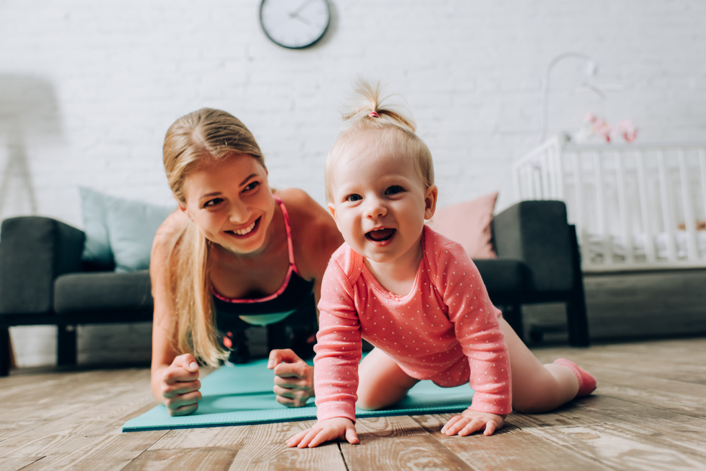 Селективный фокус ребенка, смотрящего на камеру рядом с матерью, делающей доску на фитнес-коврик дома  - Фото, изображение