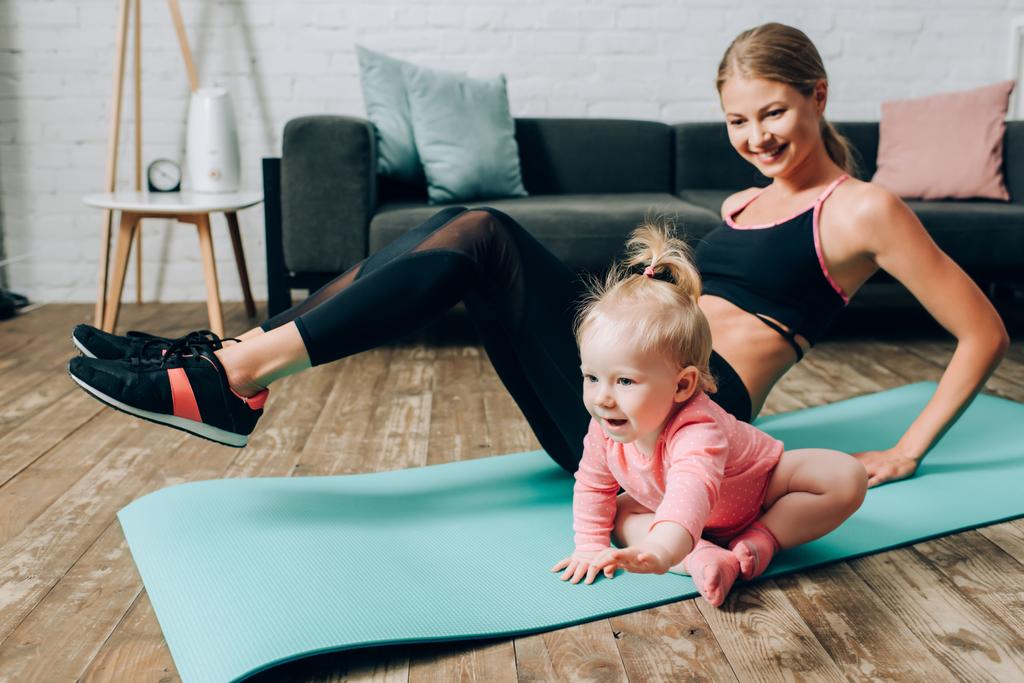 Focus selettivo della bambina seduta sul tappeto fitness mentre la madre si allena a casa   - Foto, immagini