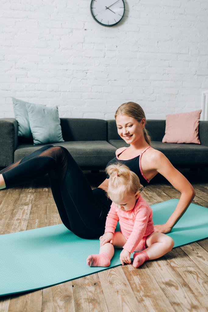 Selective focus of baby girl sitting on fitness mat while mother training at home  - Photo, Image