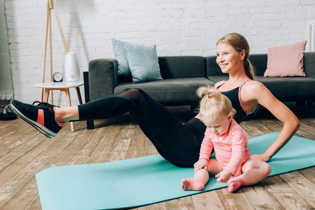 Foco seletivo do treinamento da mulher no tapete de fitness perto da filha do bebê na sala de estar  - Foto, Imagem