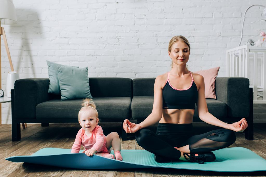 Frau sitzt in Yoga-Pose auf Fitnessmatte neben Baby-Mädchen  - Foto, Bild