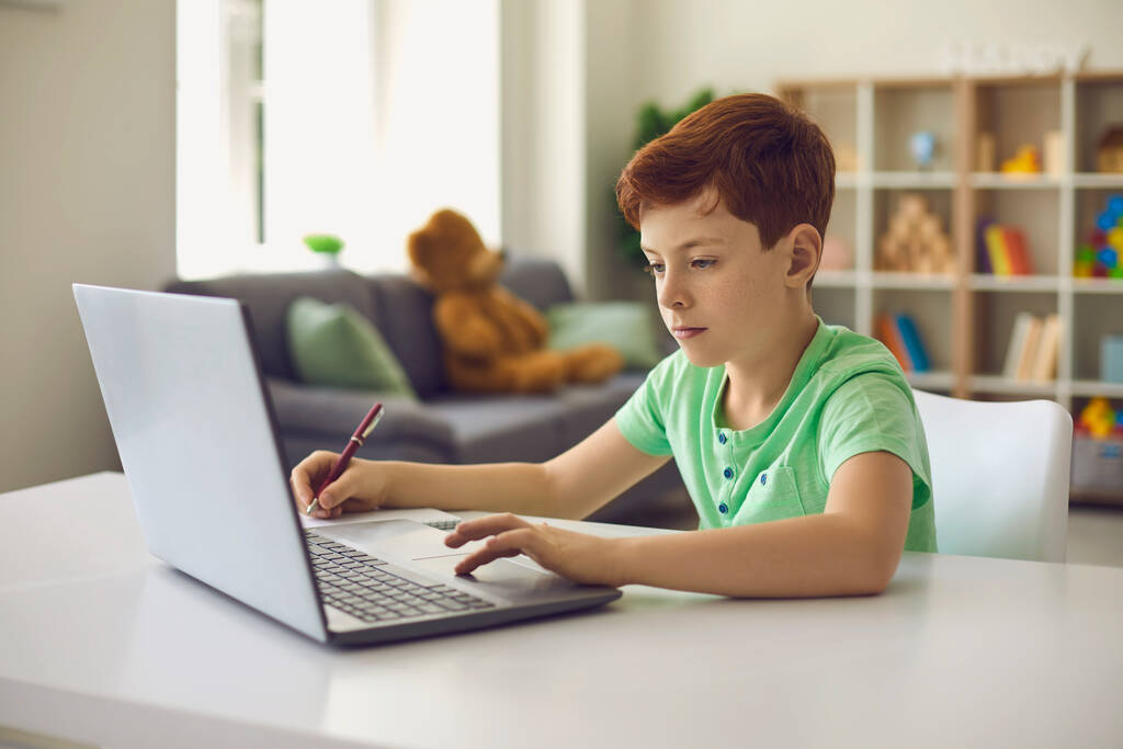 オンライン教育。ノートパソコンでレッスンを見てノートを取ることに焦点を当てた少年。家から遠隔で勉強する学童 - 写真・画像