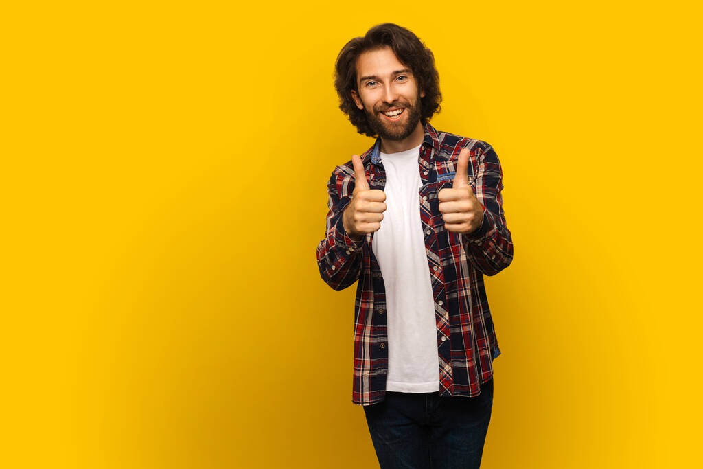 Knappe glimlachende man met krullend haar tonen duim omhoog geïsoleerd op gele achtergrond - Foto, afbeelding