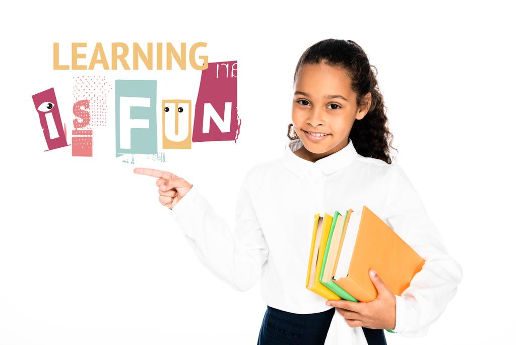 african american schoolgirl pointing with finger at learning is fun lettering while holding books isolated on white - Photo, Image