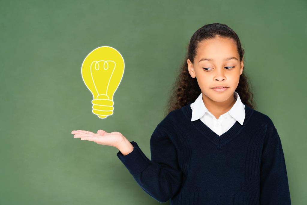 skeptická školačka ukazuje rukou na žárovku na zelené tabuli  - Fotografie, Obrázek