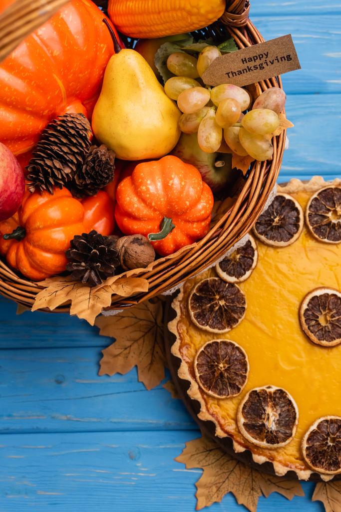 top view of autumnal harvest in wicker basket with happy thanksgiving card near pumpkin pie on blue wooden background - Photo, Image