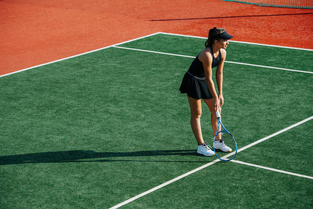 Tienermeisje in een zwarte sportieve outfit tennissen op een gloednieuwe baan, leunend op een racket tussen sets. - Foto, afbeelding