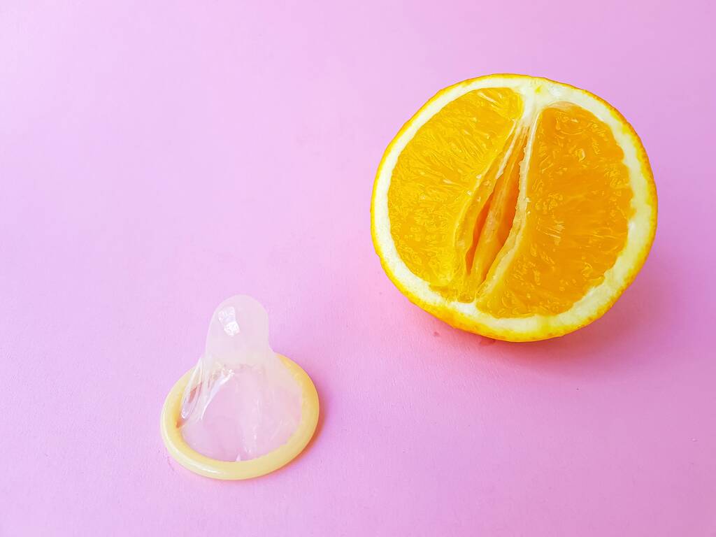 The concept of safe sex and protection. A vagina symbol. A juicy orange and a condom on a bright pink background. - Photo, image