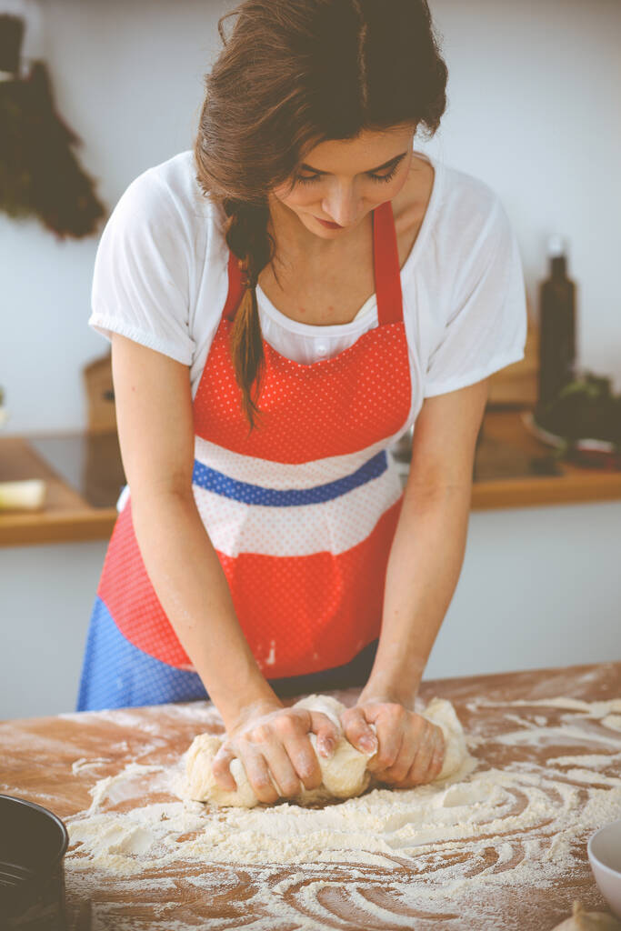 Młoda brunetka gotuje pizzę lub ręcznie robiony makaron w kuchni. Gospodyni domowa przygotowuje ciasto na drewnianym stole. Koncepcja diety, żywności i zdrowia - Zdjęcie, obraz