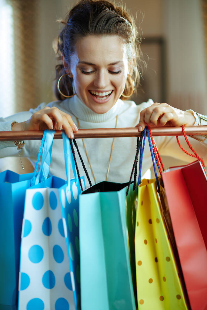 happy stylish female in white sweater and skirt looking in shopping bag hanging on copper clothes rail. - Photo, Image