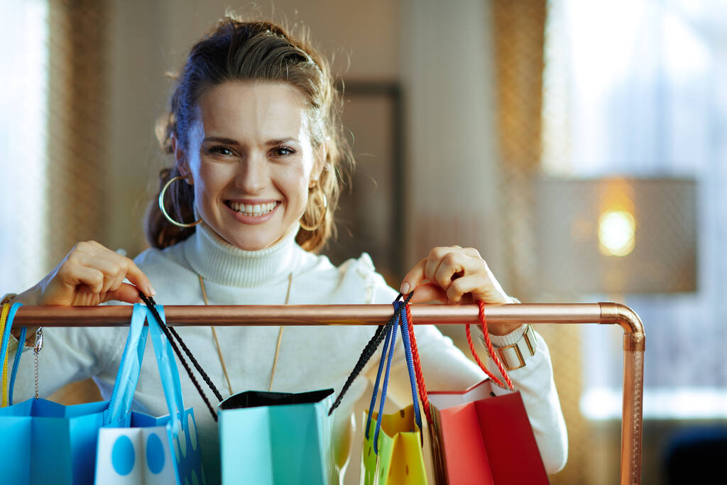 sorrindo jovem mulher em camisola branca e saia perto de sacos de compras coloridos pendurados no trilho de roupas de cobre. - Foto, Imagem