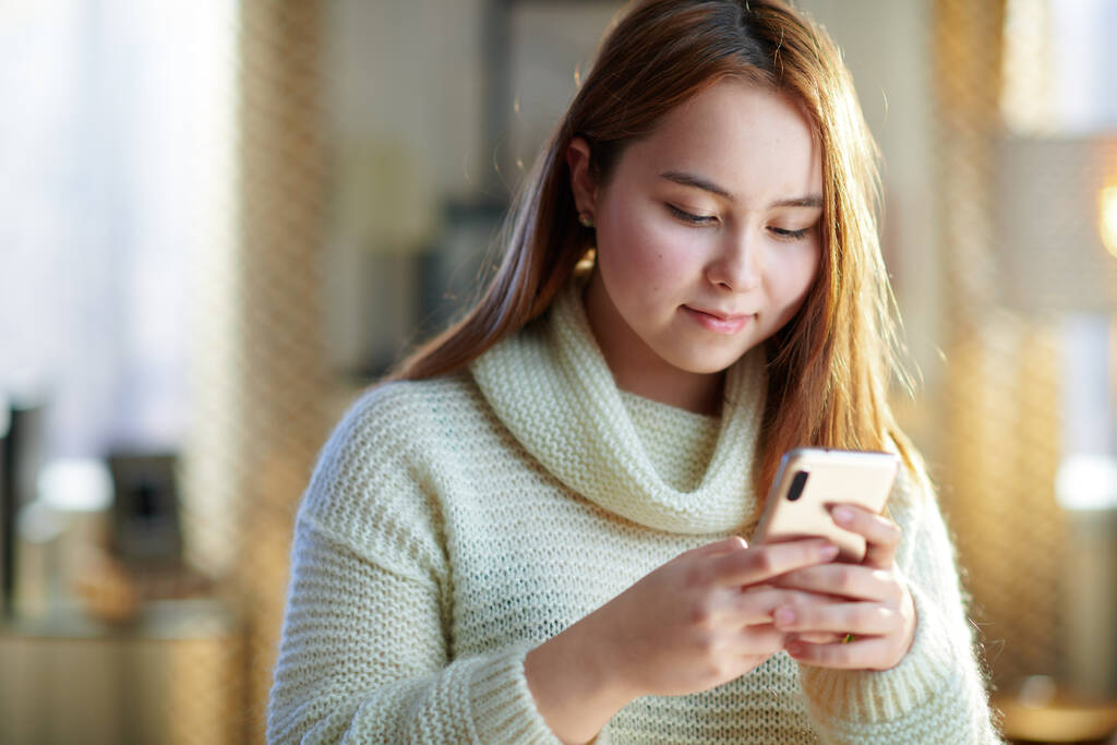 feliz chica adolescente moderna con el pelo rojo en suéter blanco en el hogar moderno en el día de invierno soleado navegar por las redes sociales en un teléfono inteligente. - Foto, imagen