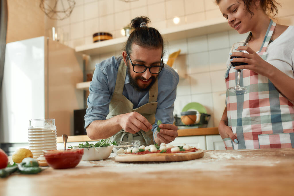 En iyi aşçı Genç çift evde birlikte pizza yapıyor. Önlüklü bir adam, kadın şarap içerken profesyonel aşçı hamura fesleğen ekliyor. Hobi, yaşam tarzı - Fotoğraf, Görsel