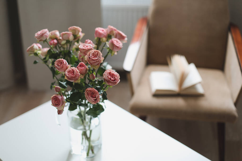Intérieur de chambre moderne avec fauteuil, livre et roses fraîches sur la table. Intérieur du beau salon décoré de fleurs - Photo, image