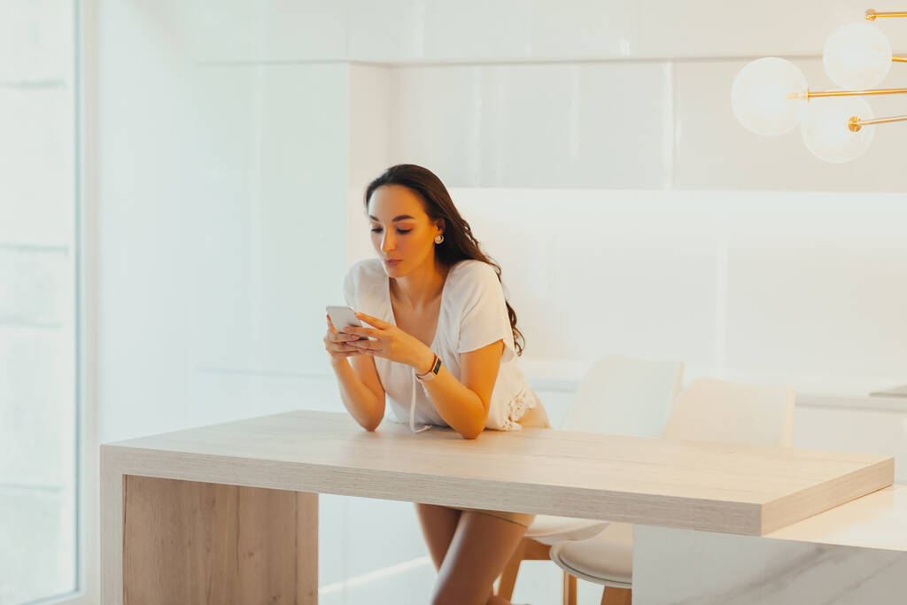 Серьёзная молодая красивая женщина пользуется телефоном в минималистичной светлой кухне. Создание и коммуникация контента. Ожидание звонка или сообщения - Фото, изображение