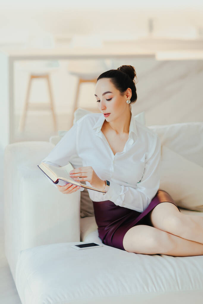 Mujer hermosa joven y seria está sentada en el sofá y leyendo libro. Interior minimalista, descanso en casa - Foto, Imagen
