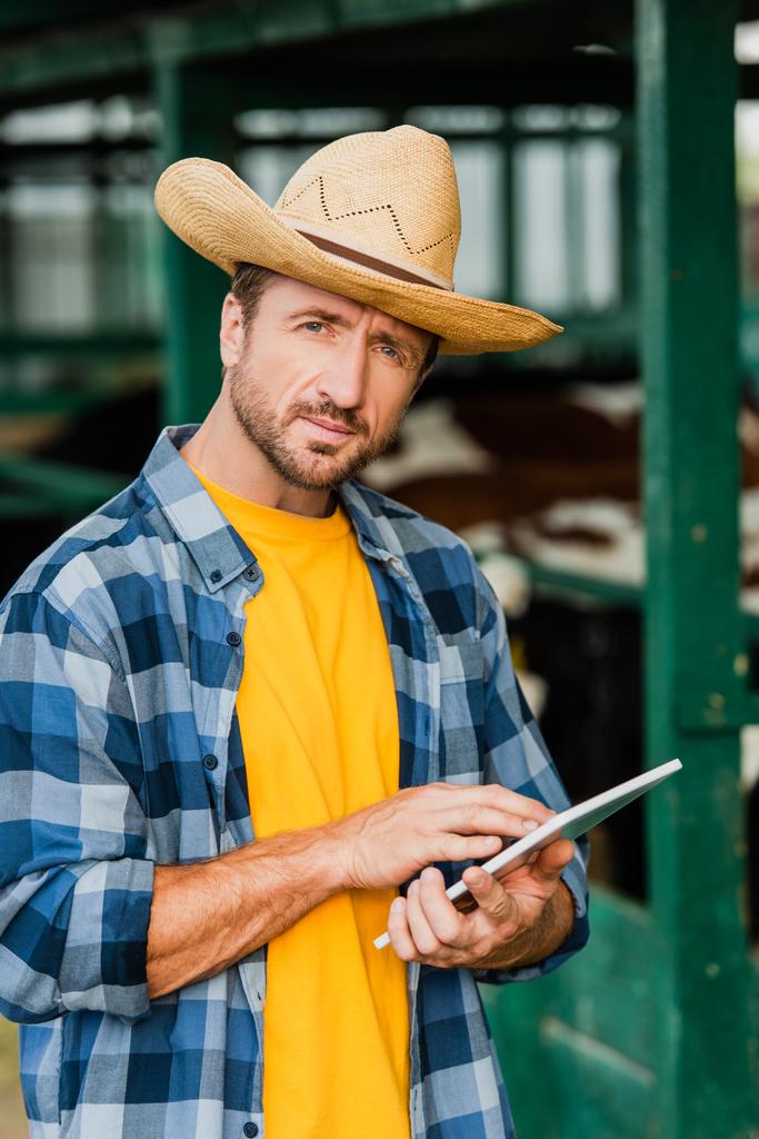 ranchero en sombrero de paja y camisa a cuadros usando tableta digital mientras mira a la cámara  - Foto, imagen