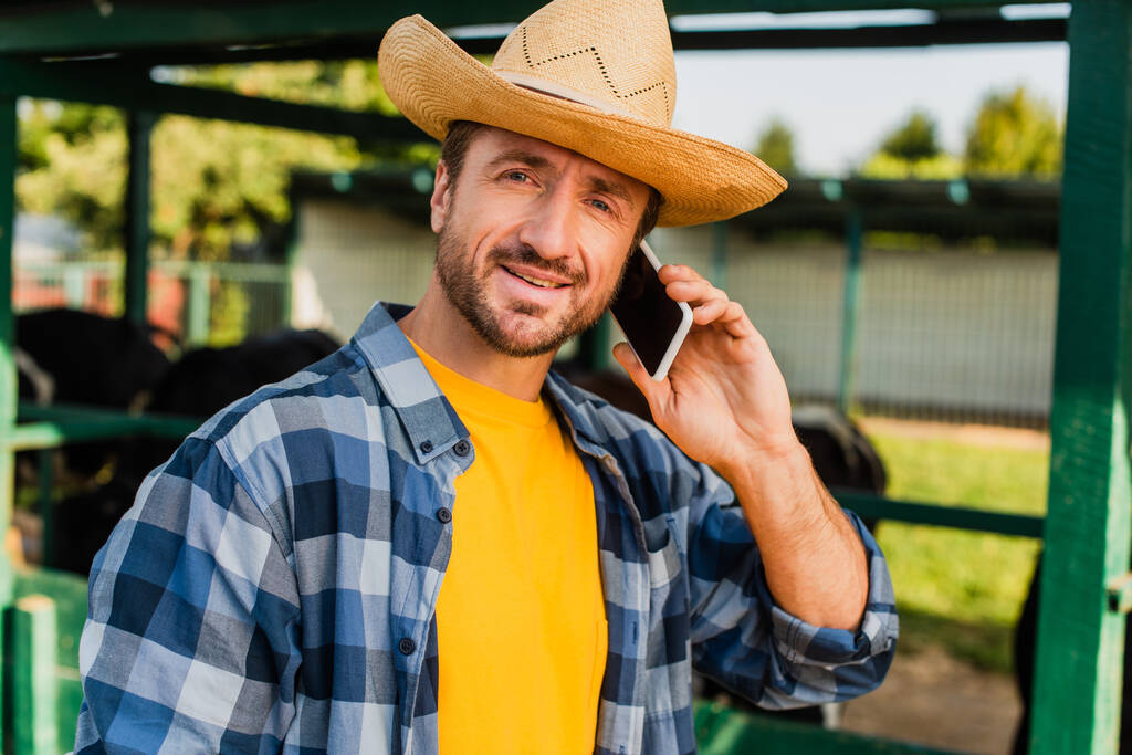 farmer kockás ing és szalma kalap beszél okostelefonon, miközben nézi a kamera  - Fotó, kép