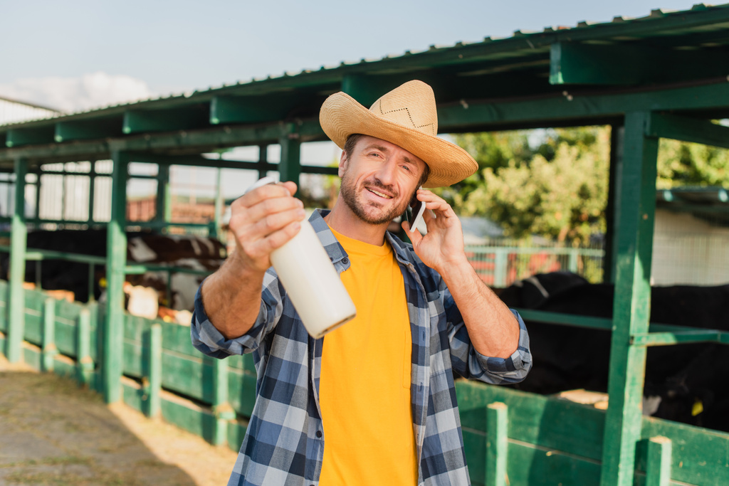 ranchero en sombrero de paja y camisa a cuadros hablando por teléfono móvil mientras muestra botella de leche fresca cerca de establo - Foto, Imagen