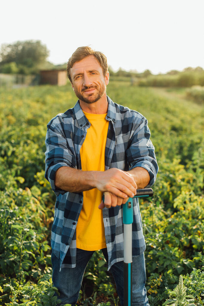 фермер в клетчатой рубашке смотрит в камеру, стоя с лопатой в поле - Фото, изображение