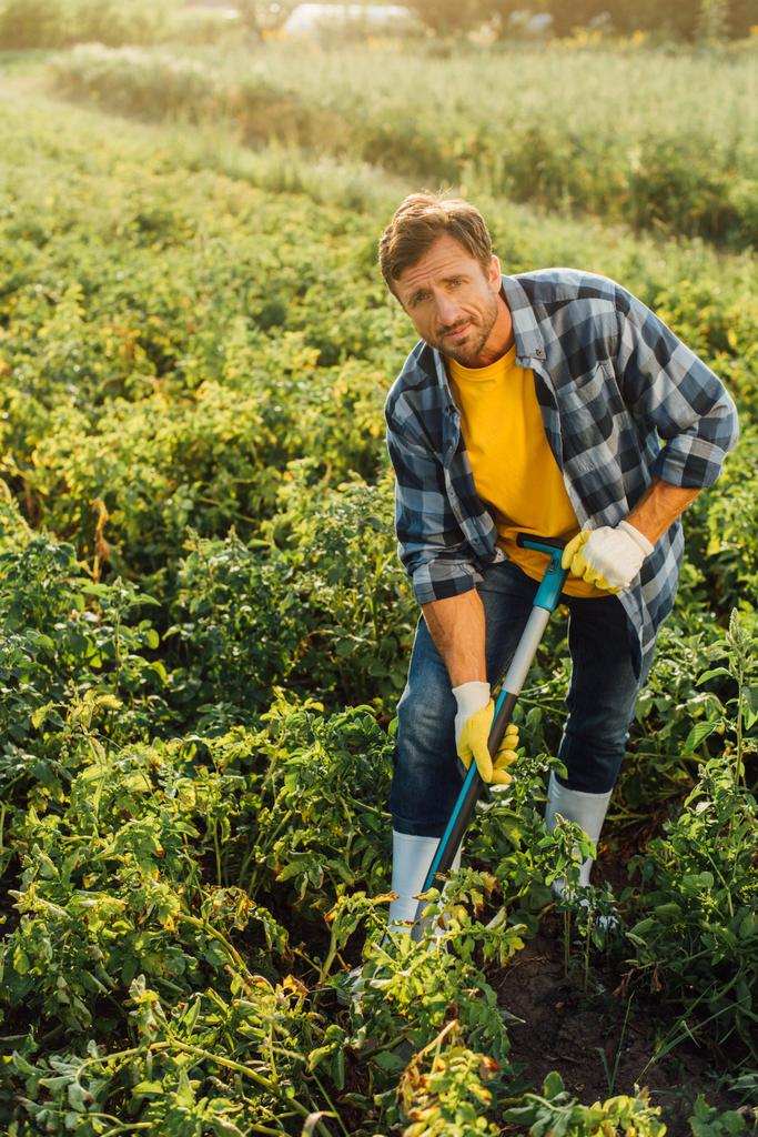 фермер в клетчатой рубашке, перчатках и резиновых сапогах копает в поле, глядя в камеру - Фото, изображение