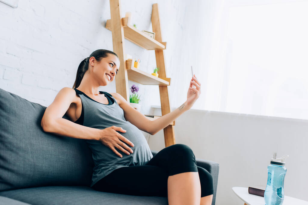 Zwangere sportvrouw neemt selfie met smartphone in de buurt van sportfles  - Foto, afbeelding