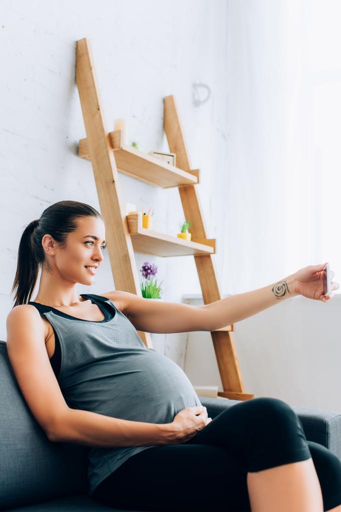 Focus sélectif de la jeune femme enceinte en tenue de sport prenant selfie avec smartphone sur canapé  - Photo, image