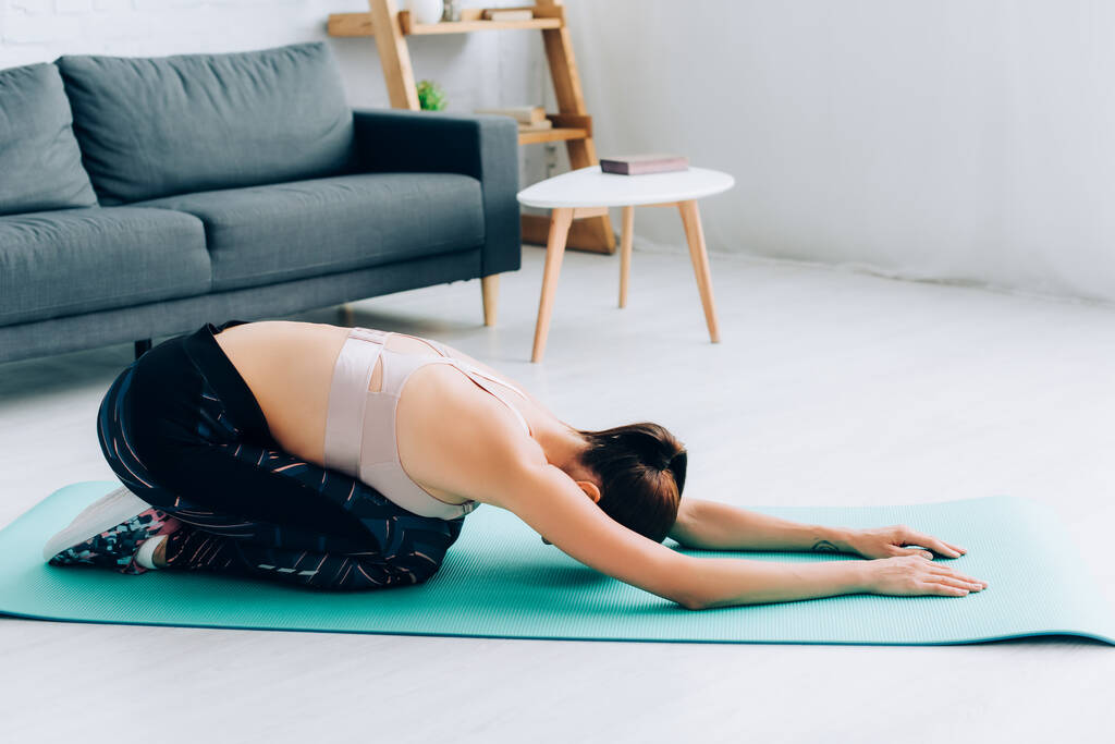 Zijaanzicht van brunette sportvrouw stretching terwijl zitten op fitness mat in de woonkamer  - Foto, afbeelding