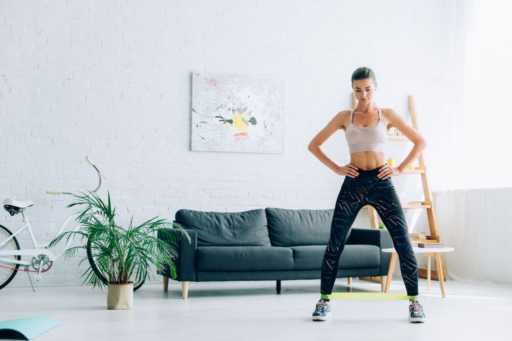 Spor giyimli ve spor ayakkabılı bir kadın evde antrenman yaparken direniş bandı kullanıyor.  - Fotoğraf, Görsel