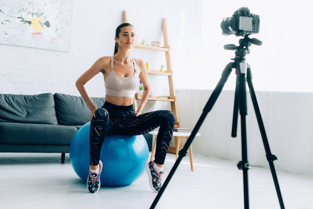 Foco seletivo de esportista sentado na bola de fitness perto da câmera digital no tripé em casa  - Foto, Imagem