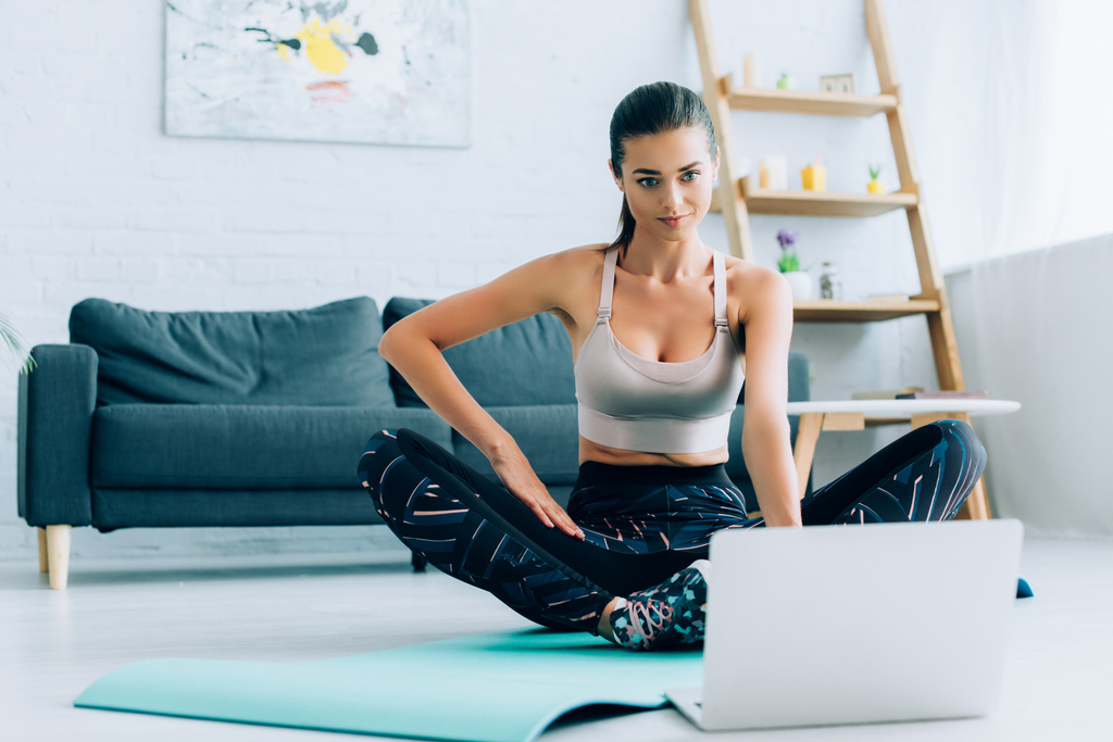 Focus selettivo della donna bruna in abbigliamento sportivo guardando l'allenamento online sul laptop sul tappeto fitness - Foto, immagini