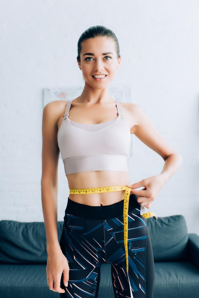 Young sportswoman looking at camera while measuring waist with tape at home  - Photo, Image