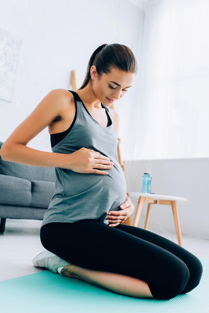 Mujer deportista embarazada tocando el vientre mientras se sienta en la alfombra de fitness en la sala de estar  - Foto, Imagen