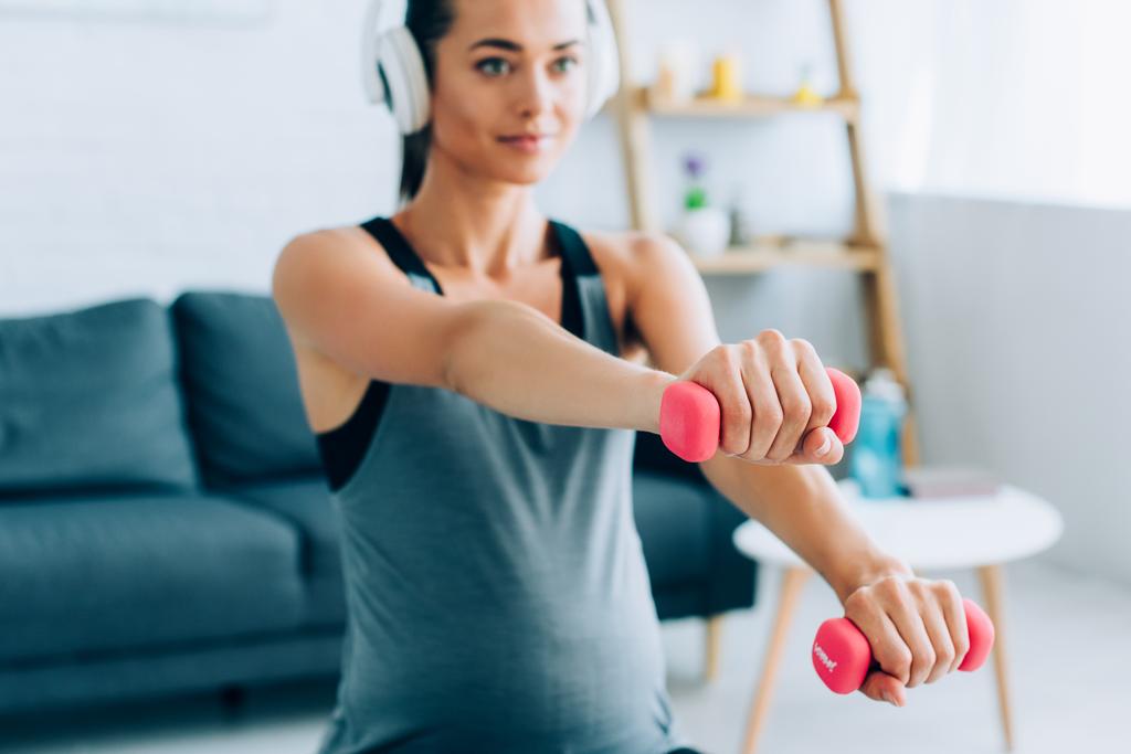 Selective focus of pregnant woman in headphones training with pink dumbbells at home  - Photo, Image