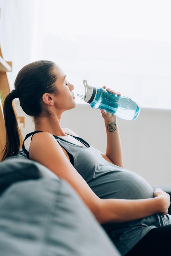 Enfoque selectivo de la deportista embarazada bebiendo agua mientras está sentada en el sofá en casa  - Foto, Imagen