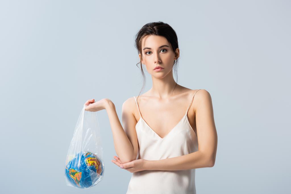 jonge vrouw in zijden jurk houden plastic zak met bol geïsoleerd op grijs, ecologie concept  - Foto, afbeelding