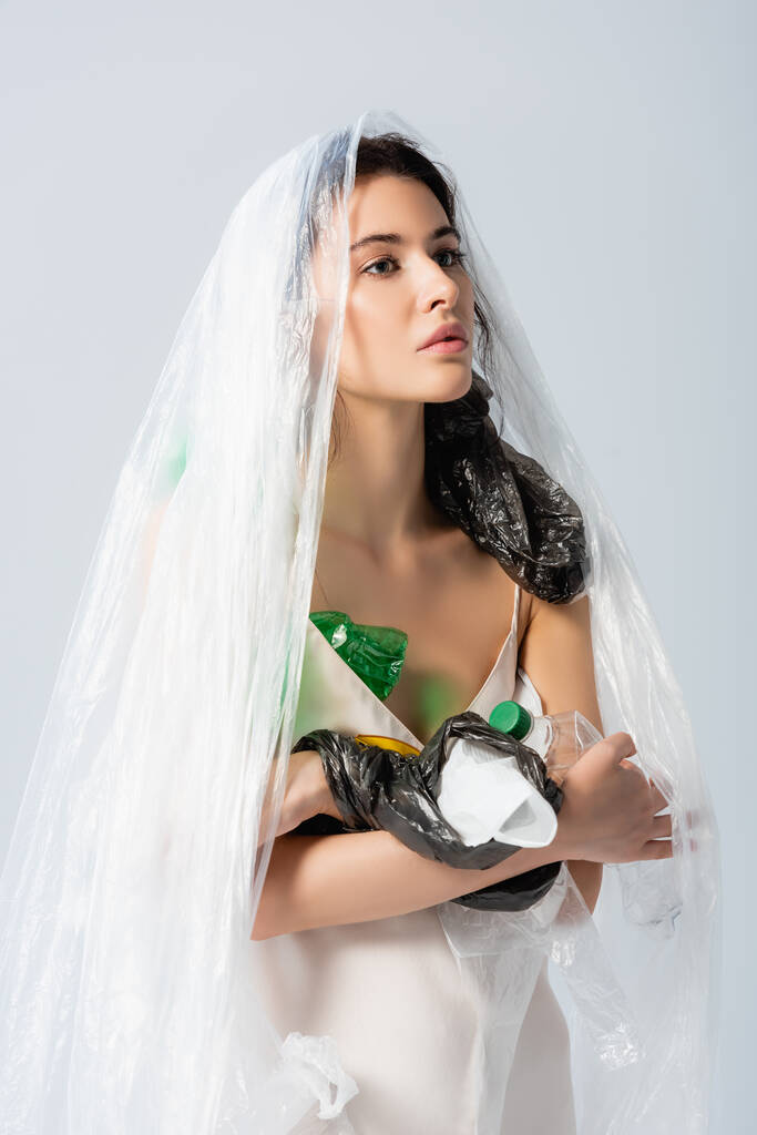 mujer morena con bolsa de plástico en la cabeza de pie en vestido de seda con botellas vacías aisladas en blanco, concepto de ecología   - Foto, Imagen