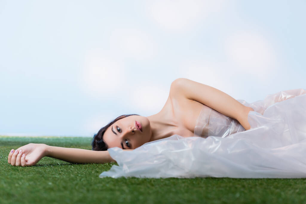 young brunette woman wrapped in polyethylene lying on green grass and looking at camera on blue, ecology concept  - Photo, Image